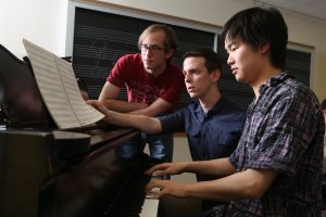 Student at piano, prfessor turning music book pages, second student leaning in 