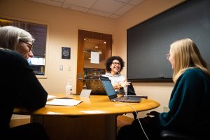 Student in background laughing and his class peer and professor are in the foreground at a round table