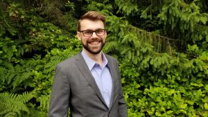 A man in a suit and glasses is smiling in front of some green trees.