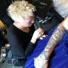 A blond woman kneels on the floor to sew adjustments into a blue dress that a model is wearing.