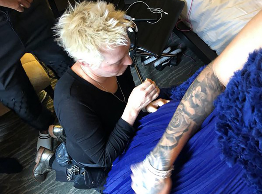A blond woman kneels on the floor to sew adjustments into a blue dress that a model is wearing.