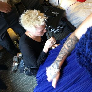 A blond woman kneels on the floor to sew adjustments into a blue dress that a model is wearing.