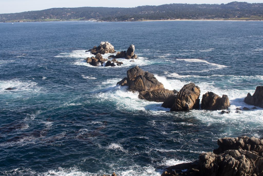 Blue ocean waters are seen swirling around rocks on the coast.