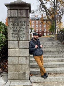 A bearded man in a baseball cap and winter coat leans against a pillar at the bottom of a set of stairs.
