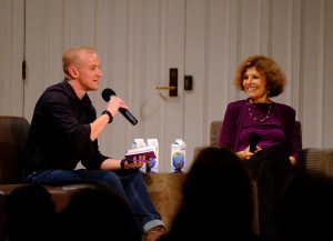 A man and a woman sit on a stage as the man speaks into a microphone.