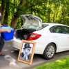 A student wearing a mask unloads a rubber bin from the trunk of his car while a portrait of Harpo leans up against it.