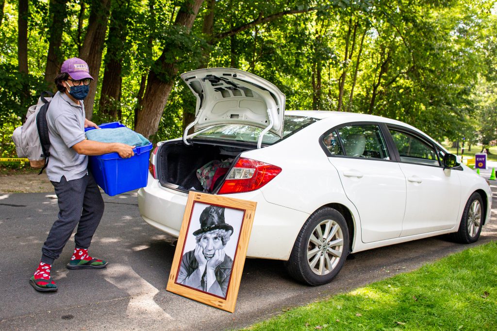 A student wearing a mask unloads a rubber bin from the trunk of his car while a portrait of Harpo leans up against it.