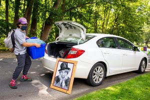 A student wearing a mask unloads a rubber bin from the trunk of his car while a portrait of Harpo leans up against it.