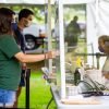 Two students check in to a temporary testing site by holding their I.D.s up to a plexiglass barrier, on the other side of which a volunteer checks them in.