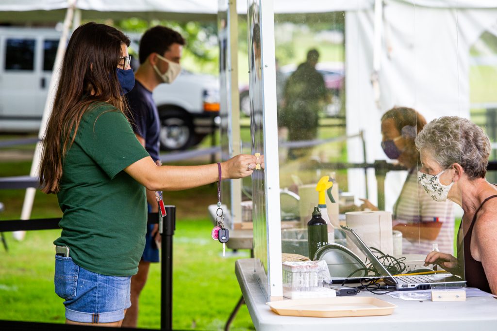 Two students check in to a temporary testing site by holding their I.D.s up to a plexiglass barrier, on the other side of which a volunteer checks them in.