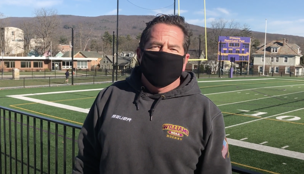 Williams College groundkeeper Jimmy Menard wears a black face mask and stands in front of the football field.